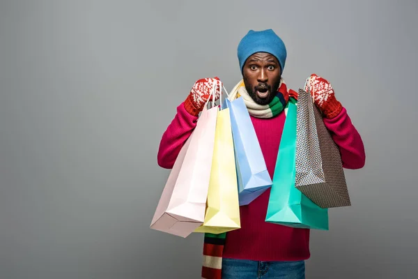 Surpris homme afro-américain en tenue d'hiver avec des sacs à provisions sur fond gris — Photo de stock