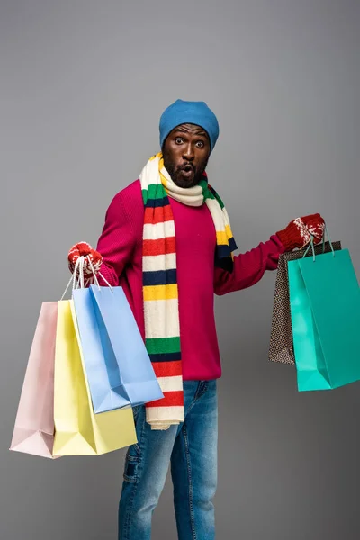 Surprised african american man in winter outfit with shopping bags on grey background — Stock Photo