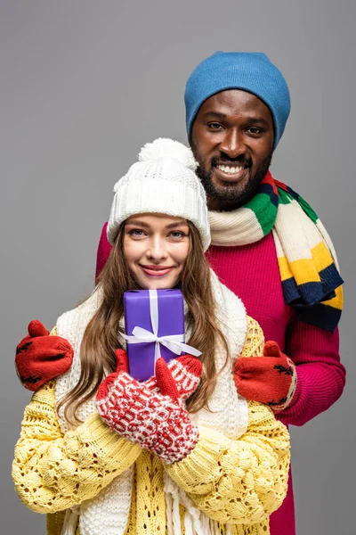 Glückliches gemischtrassiges Paar im winterlichen Outfit mit Geschenk isoliert auf grau — Stockfoto