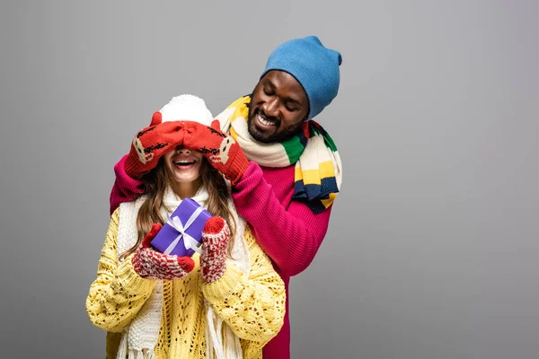 Щасливий афроамериканський чоловік в зимовому вбранні, прикриваючи очі дівчини, тримає подарунок ізольовано на сірому — стокове фото