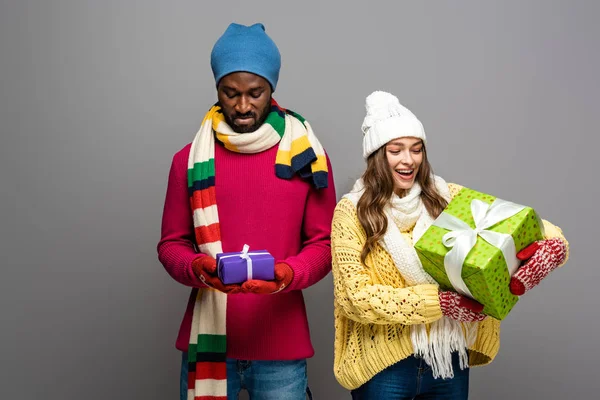 Feliz y molesto interracial pareja en invierno traje celebración regalos en gris fondo - foto de stock