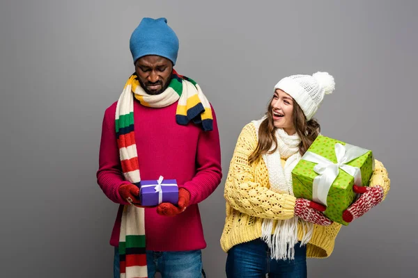 Feliz e chateado interracial casal no inverno roupa segurando presentes no fundo cinza — Fotografia de Stock