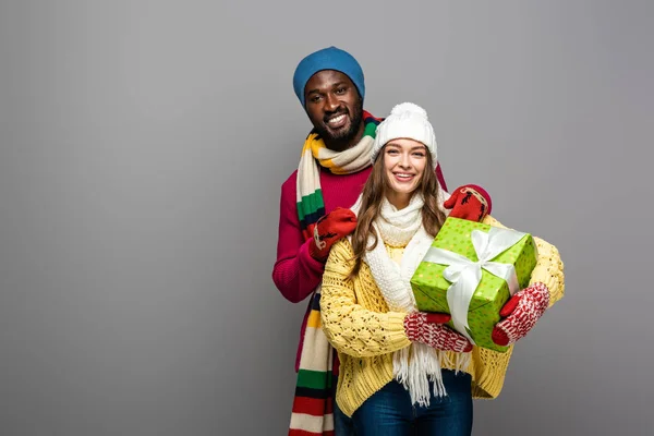 Glückliches gemischtrassiges Paar im winterlichen Outfit mit Geschenk isoliert auf grau — Stockfoto