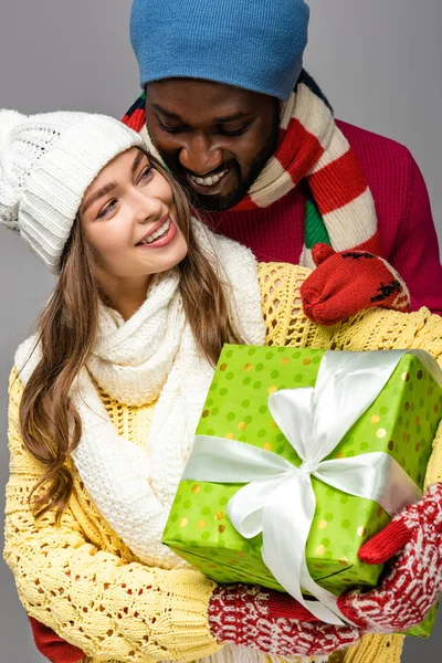 Smiling interracial couple in winter outfit holding present isolated on grey — Stock Photo