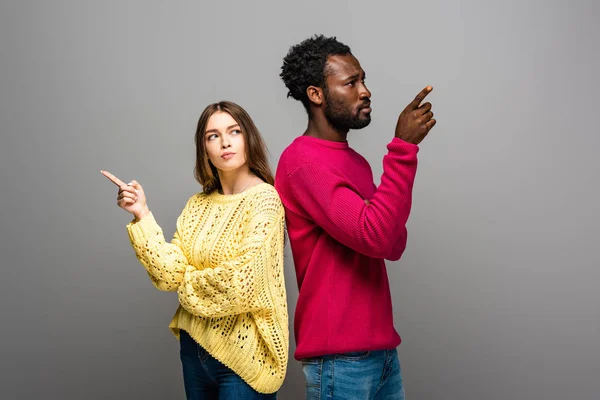 Thoughtful interracial couple in knitted sweaters standing back to back and pointing with fingers on grey background — Stock Photo