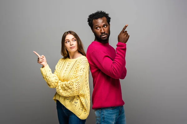Couple interracial réfléchi en chandails tricotés debout dos à dos et pointant avec les doigts sur fond gris — Photo de stock