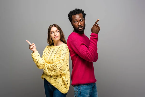 Pareja interracial reflexivo en suéteres de punto de pie espalda con espalda y señalando con los dedos sobre fondo gris - foto de stock