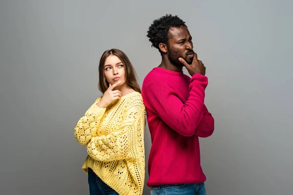 Вдумчивая межрасовая пара в вязаных свитерах, стоящая спиной к спине на сером фоне — стоковое фото