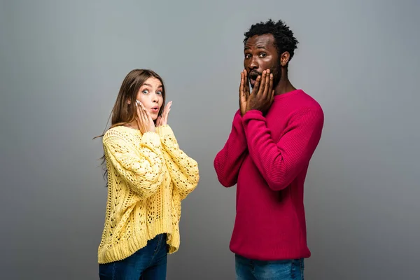 Couple interracial choqué en chandails tricotés avec les mains près du visage sur fond gris — Photo de stock