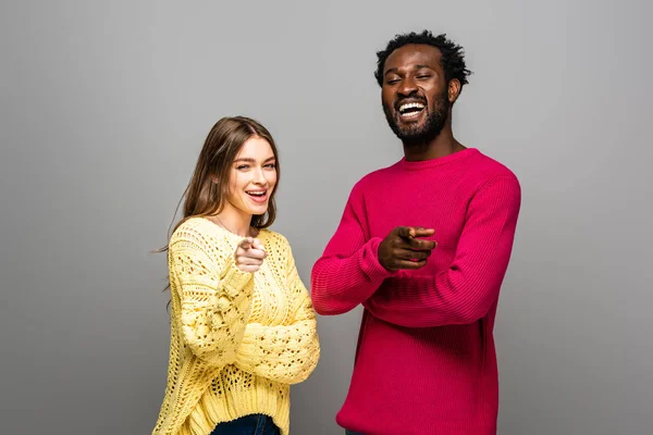 Усміхнена міжрасова пара в трикотажних светрах, що вказують пальцями на сірому фоні — стокове фото