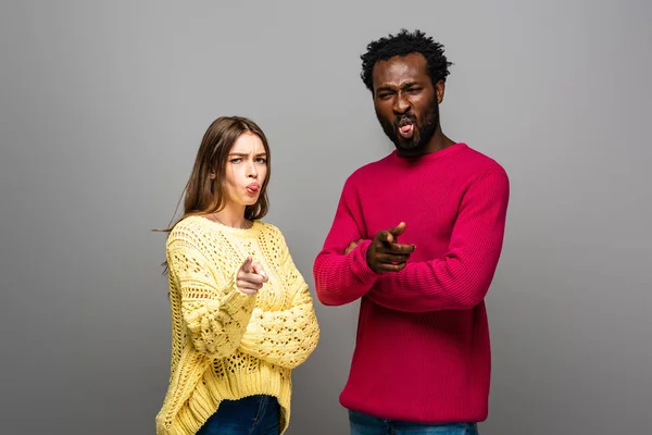 Interracial couple in knitted sweaters pointing with fingers and showing tongues on grey background — Stock Photo