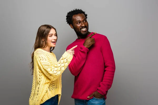 Lächelndes gemischtrassiges Paar in Strickpullovern, die mit Fingern auf grauem Hintergrund zeigen — Stockfoto