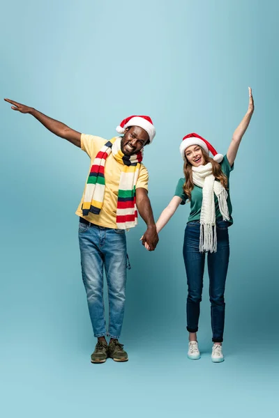 Feliz interracial pareja en santa sombreros y bufandas cogidos de la mano sobre fondo azul - foto de stock