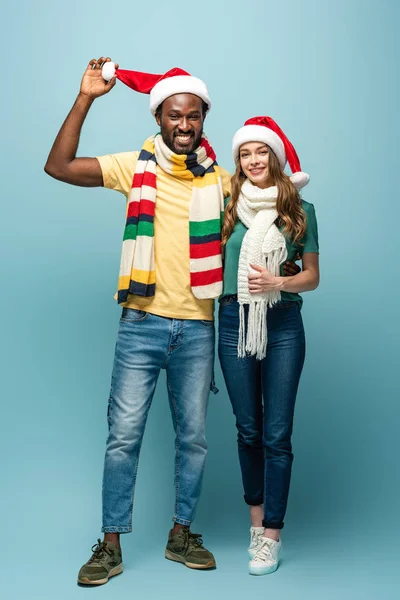 Feliz pareja interracial en sombreros de santa y bufandas sobre fondo azul - foto de stock