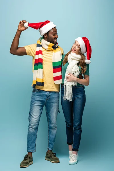Feliz pareja interracial en sombreros de santa y bufandas sobre fondo azul - foto de stock