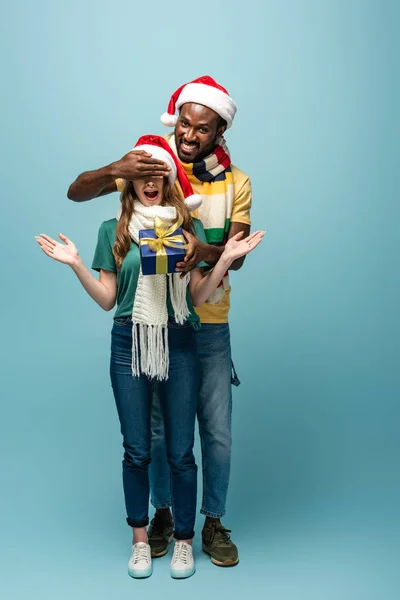 Glücklicher afrikanisch-amerikanischer Mann mit Weihnachtsmütze und Schal, der seiner überraschten Freundin ein Geschenk macht und die Augen vor blauem Hintergrund verhüllt — Stockfoto
