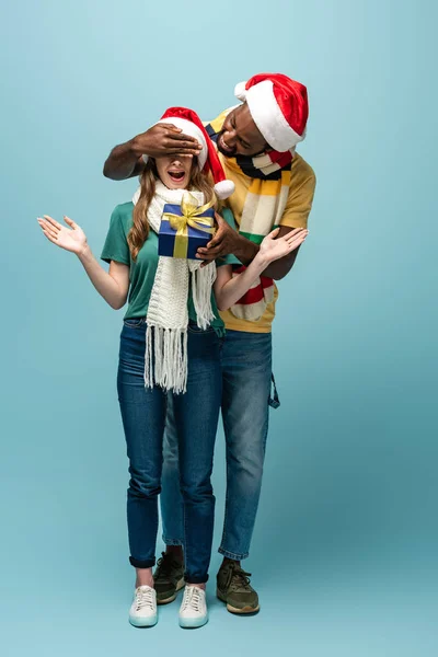 Feliz africano americano hombre en santa sombrero y bufanda regalo a la novia sorprendida y cubriendo los ojos sobre fondo azul - foto de stock