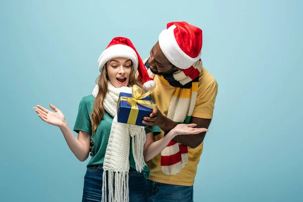 Feliz africano americano hombre en santa hat y bufanda regalo a sorprendido novia aislado en azul - foto de stock