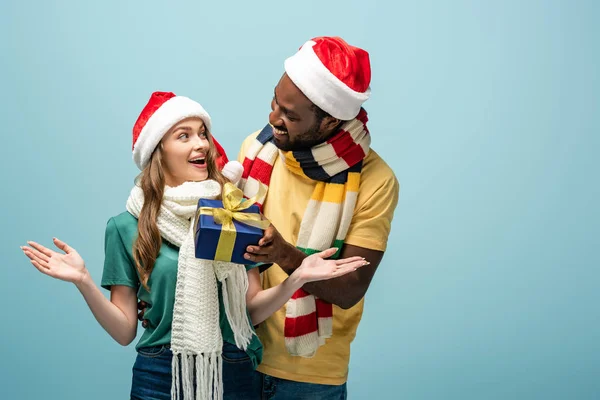 Glücklich afrikanisch-amerikanischer Mann in Weihnachtsmütze und Schal Geschenk an überraschte Freundin isoliert auf blau — Stockfoto