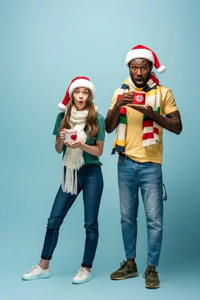 Shocked interracial couple in santa hats and scarves holding mugs with cocoa on blue background — Stock Photo