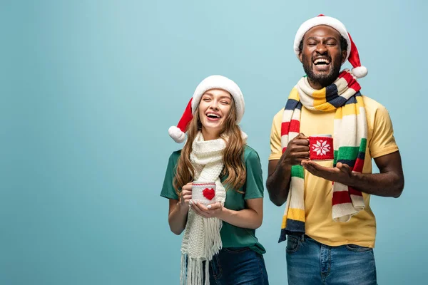 Laughing interracial couple in santa hats and scarves holding mugs with cocoa isolated on blue — Stock Photo