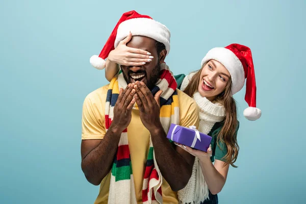 Happy girl in santa hat and scarf covering eyes on african american boyfriend and gifting present isolated on blue — Stock Photo