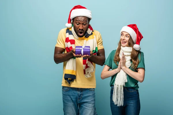 Happy girl in santa hat and scarf gifting present to african american shocked boyfriend isolated on blue — Stock Photo