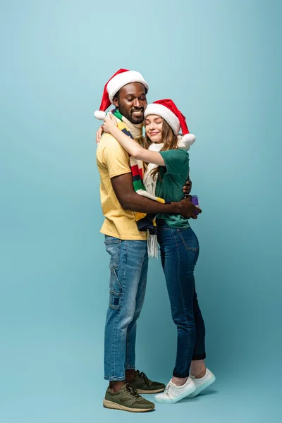 Glückliches interracial Paar, das sich mit Geschenk auf blauem Hintergrund umarmt — Stockfoto