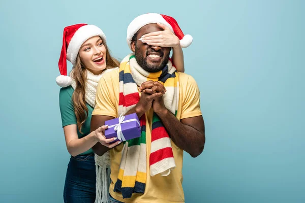 Щаслива дівчина в капелюсі Санта і шарфі, що закриває очі на афроамериканського американського хлопця і подарунок ізольовано на синьому — стокове фото