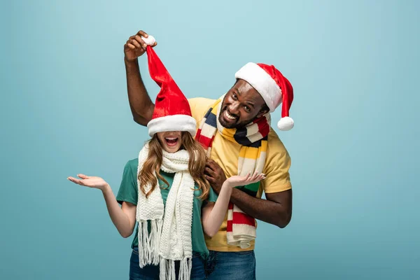Felice coppia interrazziale in cappelli di Babbo Natale e sciarpe divertirsi isolato su blu — Foto stock