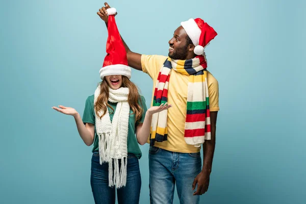 Happy interracial couple in santa hats and scarves having fun isolated on blue — Stock Photo