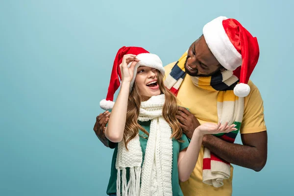 Heureux couple interracial dans santa chapeaux et foulards avoir amusant isolé sur bleu — Photo de stock