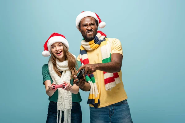 KYIV, UKRAINE - 22 AOÛT 2019 : couple interracial souriant dans des chapeaux de Père Noël et des foulards tenant des joysticks isolés sur bleu — Photo de stock