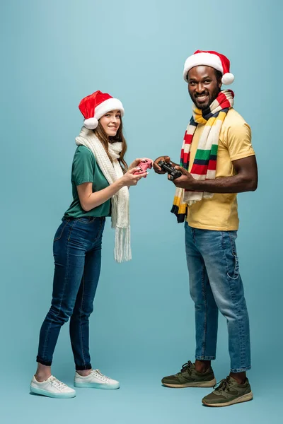 KYIV, UKRAINE - AUGUST 22, 2019: smiling interracial couple in santa hats and scarves holding joysticks — Stock Photo