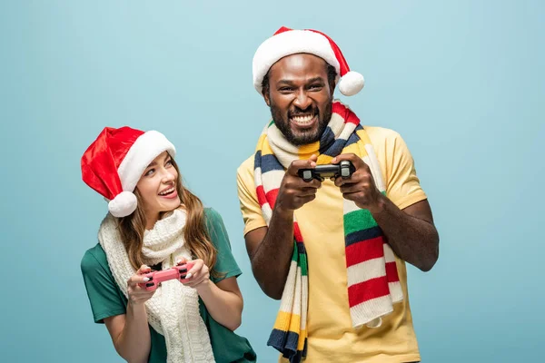 KYIV, UCRANIA - 22 de agosto de 2019: pareja interracial excitada en sombreros y bufandas de Santa Claus sosteniendo joysticks aislados en azul - foto de stock