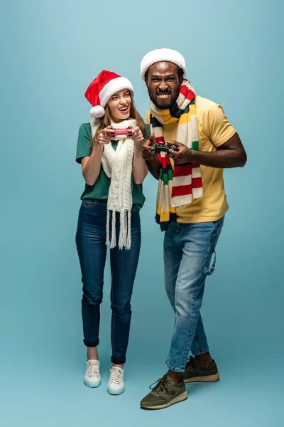 KYIV, UKRAINE - 22 AOÛT 2019 : couple interracial excité en santa chapeaux et foulards tenant des joysticks — Photo de stock