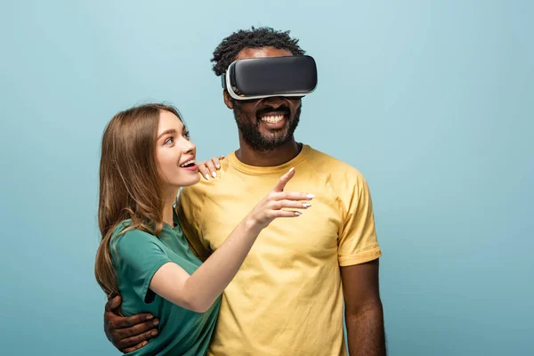 Glücklich Mädchen zeigt mit dem Finger weg in der Nähe afrikanisch-amerikanischen Freund in vr Headset auf blauem Hintergrund — Stockfoto