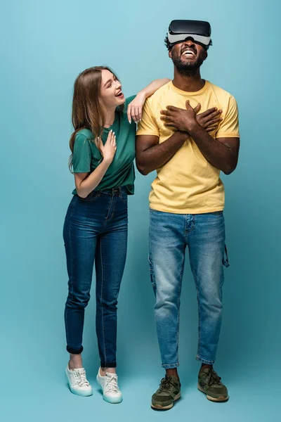 Chica feliz de pie cerca de novio afroamericano en auriculares vr sobre fondo azul - foto de stock