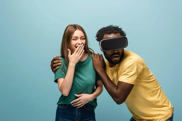 Laughing girl standing near scared african american boyfriend in vr headset on blue background — Stock Photo