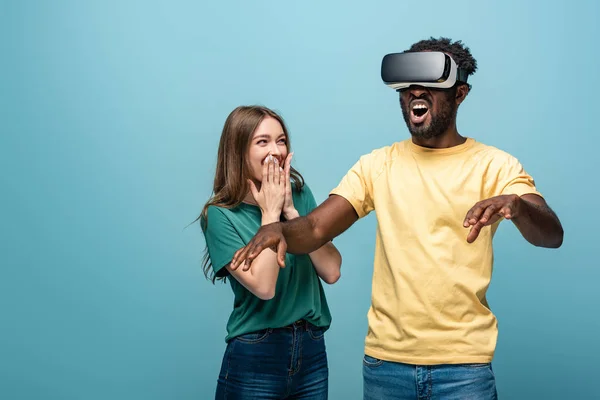 Laughing girl standing near gesturing african american boyfriend in vr headset on blue background — Stock Photo