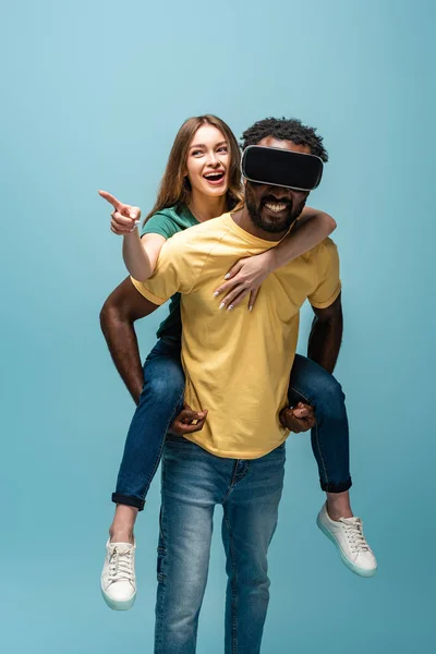 Feliz chica apuntando con el dedo mientras que piggybacking en afroamericano novio en vr auriculares aislados en azul - foto de stock