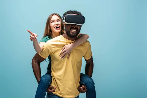 Felice ragazza che punta con il dito mentre cavalcando sul fidanzato africano americano in auricolare vr su sfondo blu — Foto stock