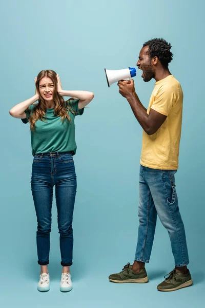 Angry african american man screaming in megaphone at girlfriend covering ears with hands on blue background — Stock Photo
