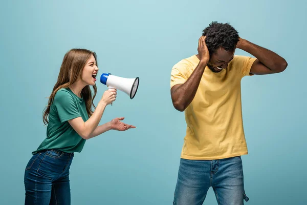 Angry girl screaming in megaphone at african american boyfriend covering ears with hands on blue background — Stock Photo