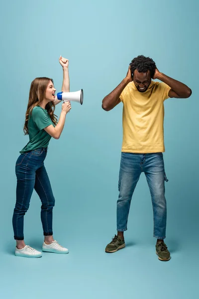 Сердитая девушка кричит в мегафоне на африканского американского бойфренда прикрывая уши руками на синем фоне — стоковое фото