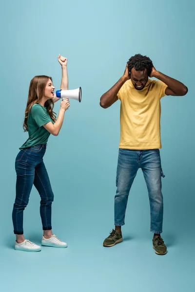 Angry girl screaming in megaphone at african american boyfriend covering ears with hands on blue background — Stock Photo