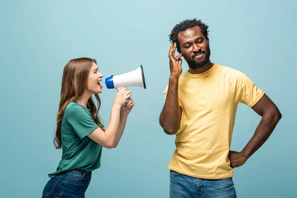 Сердитая девушка кричит в мегафоне на африканского американского бойфренда на синем фоне — стоковое фото