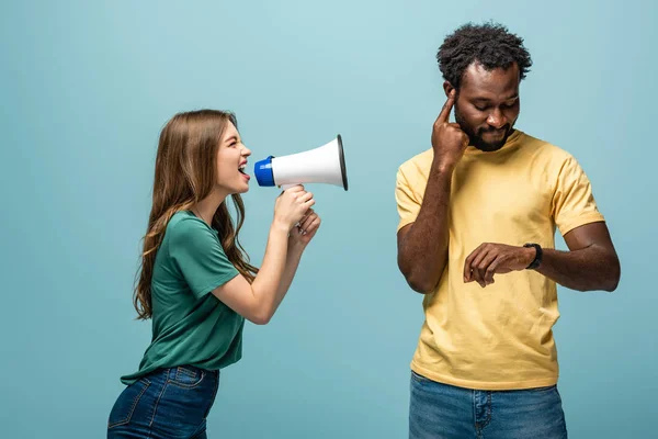 Angry girl screaming in megaphone at african american boyfriend looking at wristwatch on blue background — Stock Photo