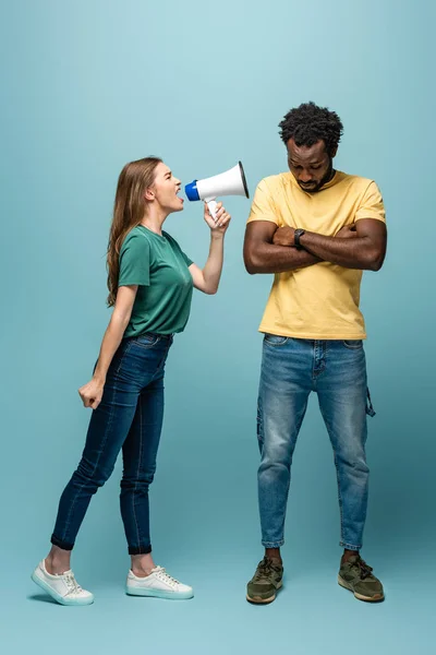 Angry girl screaming in megaphone at african american boyfriend with crossed arms on blue background — Stock Photo