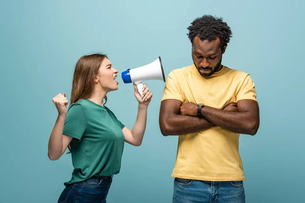Angry girl screaming in megaphone at african american boyfriend with crossed arms on blue background — Stock Photo